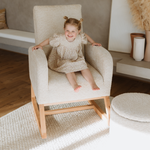 A young girl with two buns sits smiling in a boucle Myla Nursing Chair with a light wood frame, in a bright, modern living room setting.
