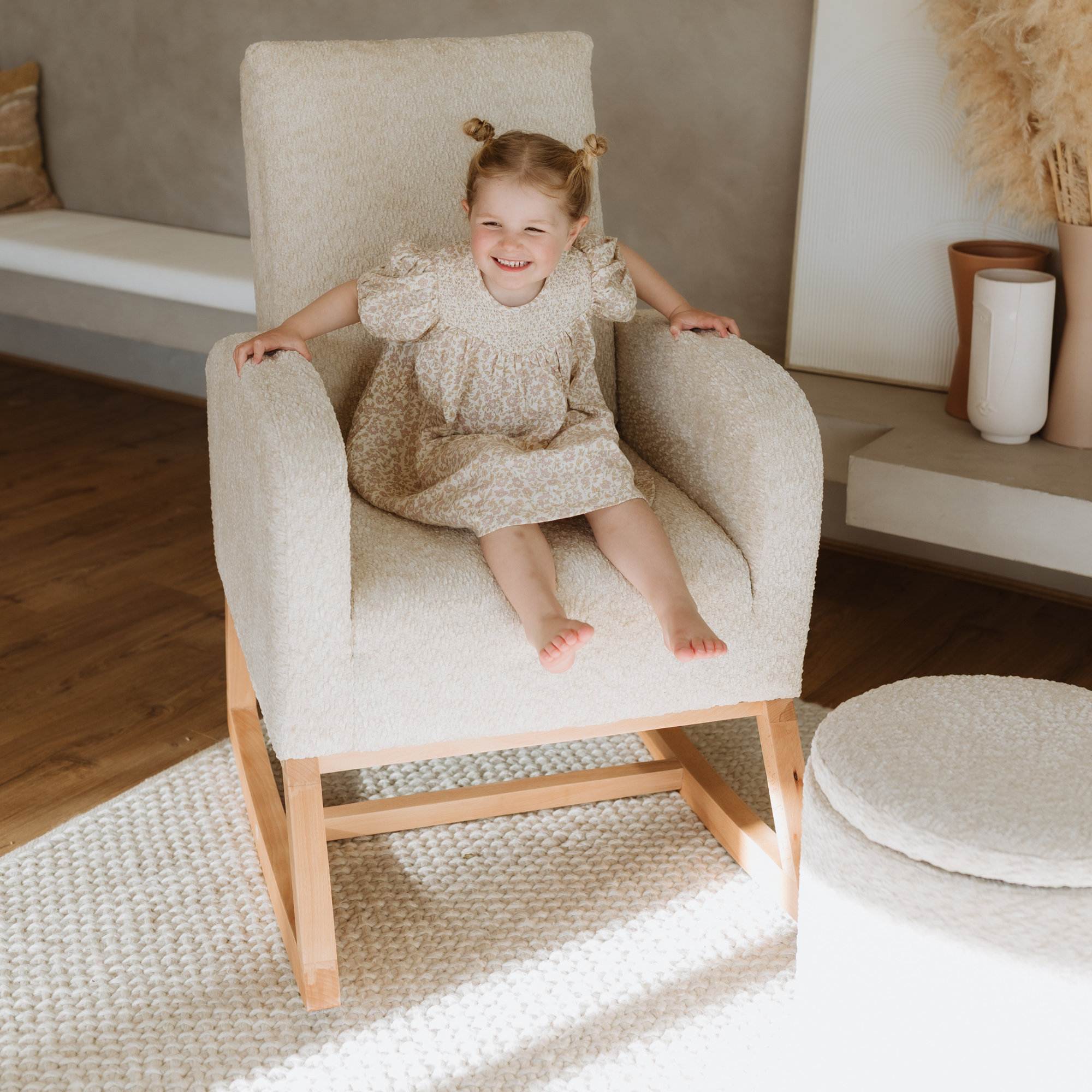 A young girl with two buns sits smiling in a boucle Myla Nursing Chair with a light wood frame, in a bright, modern living room setting.