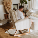 A man sits in a cream bouclé Myla rocking chair reading a book in a bright, modern living room with a fireplace and pampas grass decor.
