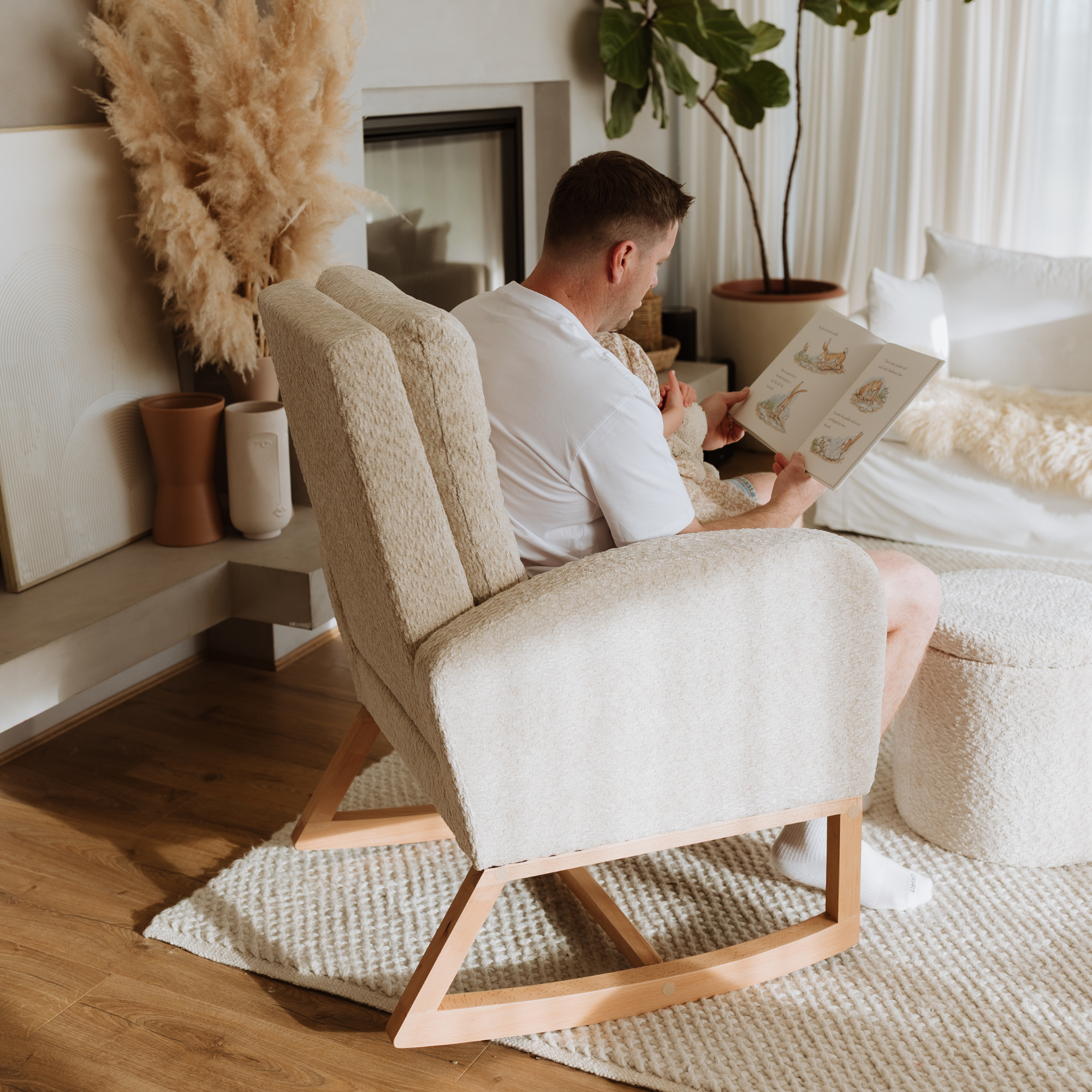 A man sits in a cream bouclé Myla rocking chair reading a book in a bright, modern living room with a fireplace and pampas grass decor.