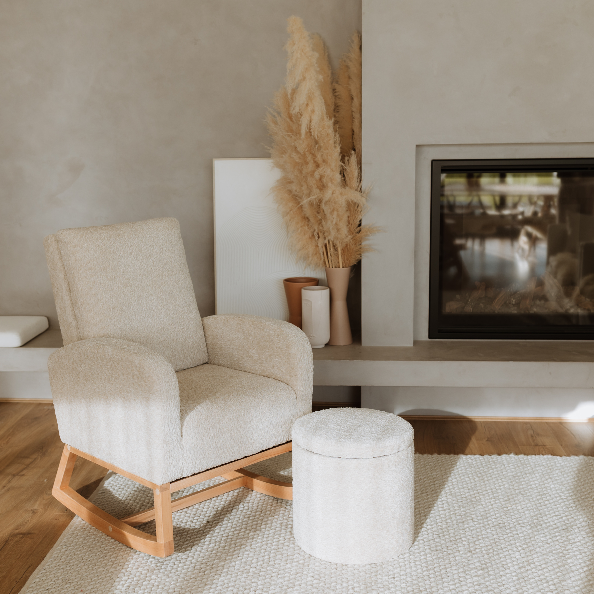 A cream-colored textured fabric rocking chair with a light wood base, styled in a minimalist living room next to a matching round ottoman, with pampas grass and a fireplace in the background.