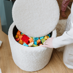 A round, light gray textured fabric Mylo ottoman with its lid propped open, filled with colorful toy building blocks. A person's hand is resting on the edge of the ottoman, and another person's legs are visible in the background on a wooden floor.