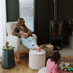 A woman relaxes in a cream upholstered Myla Nursing Chair with a matching boucle Mylo ottoman in a modern living room. A black wood-burning stove stands against a dark wall, and a child in a pink and white striped shirt sits on the floor playing with toys.