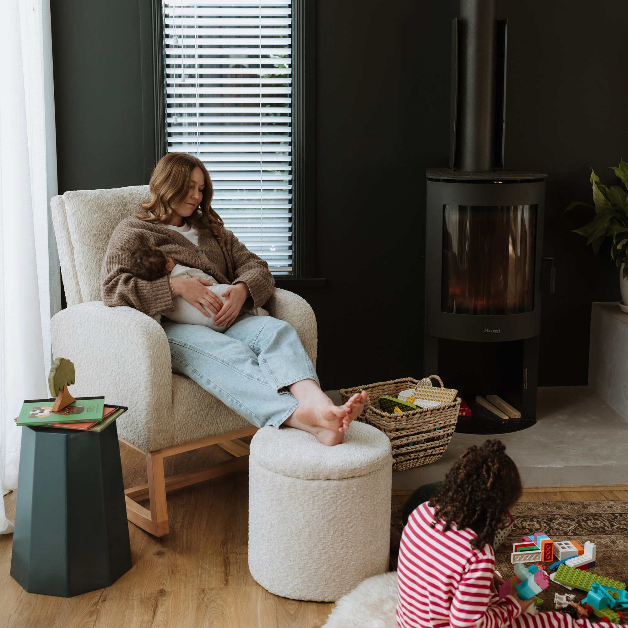 A woman relaxes in a cream upholstered Myla Nursing Chair with a matching boucle Mylo ottoman in a modern living room. A black wood-burning stove stands against a dark wall, and a child in a pink and white striped shirt sits on the floor playing with toys.