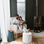 A man and a young girl sit together in a Myla chair and Mylo Ottoman, reading a green book in a modern living room with dark walls, a window with blinds, a side table, and a black wood-burning fireplace.